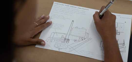 Mãos femininas sobre uma folha de papel A3 branca contendo um esboço arquitetônico em perspectiva de um conjunto de edificações, com indicações textuais manuscritas ao redor do desenho. A mão direita segura uma caneta enquanto trabalha no detalhamento da maquete técnica.