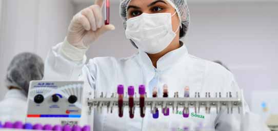 Profissional de laboratório feminina, vestindo jaleco branco, touca descartável e máscara facial, segura com a mão enluvada um tubo de ensaio com amostra de sangue contra a luz para análise. Na bancada à frente, um rack com múltiplos tubos de diferentes cores (roxo, vermelho, amarelo) e um equipamento analisador laboratorial.