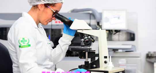 Estudante de Biomedicina, com jaleco branco com brasão institucional verde, touca descartável branca, óculos de proteção e luvas azuis, observa atentamente pelo ocular de um microscópio óptico binocular branco em laboratório de análises. Na bancada à frente, tubos de amostras coloridos em rosa e azul estão dispostos. Ao fundo, equipamentos laboratoriais compõem o ambiente científico.