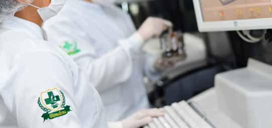 Profissional de Biomedicina em jaleco branco com brasão institucional verde — cruz médica com folhas de louro — touca descartável e luvas, opera equipamento analisador laboratorial automatizado conectado a monitor de computador. Tubos de amostras biológicas são visíveis no equipamento. A imagem mostra o trabalho técnico em laboratório de análises clínicas automatizadas.