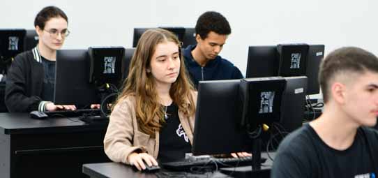 Laboratório de informática universitário com fileiras de computadores desktop de monitor preto. Em primeiro plano, uma jovem mulher de cabelo comprido castanho e suéter bege usa o mouse com concentração. Ao fundo, outros estudantes trabalham nas máquinas, incluindo um jovem negro de moletom escuro.
