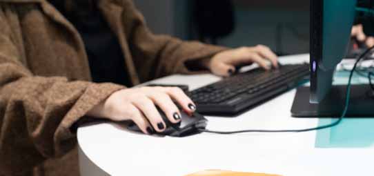 Close nas mãos de uma pessoa usando um mouse preto com fio sobre uma mesa branca, com um teclado preto e monitor ao lado. O foco nas mãos destaca a interação com equipamento de informática em ambiente de trabalho ou laboratório de dados.