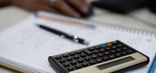 Calculadora científica preta e cinza sobre um caderno de folhas pautadas abertas, ao lado de uma caneta esferográfica preta. Ao fundo levemente desfocado, uma mão segura um mouse. A composição remete ao cotidiano de estudos em Ciências Contábeis.