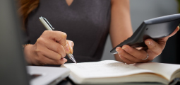 Close-up focado nas mãos de uma mulher vestindo uma blusa cinza sem mangas trabalhando em uma mesa de escritório. Ela segura uma calculadora preta com a mão esquerda e, com a mão direita, utiliza uma caneta prata para escrever anotações em um caderno de páginas brancas aberto. Em primeiro plano, no canto esquerdo, a borda de um notebook aparece levemente desfocada.