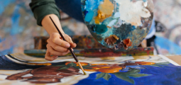 Fotografia em close-up de uma pessoa pintando uma tela. A mão em foco segura um pincel de cerdas finas, aplicando tinta em uma pintura de natureza-morta que retrata laranjas, folhas verdes e um tecido azul vibrante. Ao fundo, ligeiramente desfocada, a outra mão da pessoa segura uma paleta de madeira redonda, coberta por várias manchas grossas de tinta a óleo colorida.