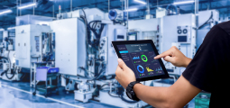 Fotografia mostrando, em primeiro plano, as mãos de uma pessoa segurando um tablet preto. A tela do dispositivo exibe um painel de controle escuro com gráficos coloridos (dashboard), incluindo um gráfico de rosca verde e azul e gráficos de barras. Ao fundo, desfocado, está o interior de uma fábrica moderna, limpa e bem iluminada, com grandes máquinas industriais CNC na cor branca e detalhes azuis.