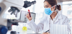 Fotografia de uma mulher trabalhando em um laboratório iluminado. Ela tem pele clara, cabelos castanhos presos, e usa um jaleco branco sobre uma camisa social e uma máscara cirúrgica azul. Ela segura um tubo de ensaio contendo um líquido vermelho escuro e o observa com atenção. Em primeiro plano, desfocado no lado esquerdo, aparece a estrutura de um microscópio preto. Sobre a bancada branca, há também um pequeno frasco de vidro contendo um líquido amarelo.