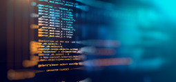 Uma imagem em close-up e com foco seletivo de uma tela escura preenchida por linhas de código de programação de computador em fontes coloridas (laranja, amarelo e azul-claro). O lado esquerdo mostra o texto do código com clareza, revelando comandos lógicos e booleanos, enquanto o lado direito se dissolve de forma abstrata em um forte brilho de luz azul-turquesa difusa com efeito de desfoque (bokeh), sugerindo um ambiente cibernético.