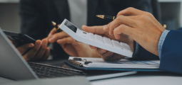 Close-up das mãos de dois profissionais trabalhando juntos em uma mesa de escritório. Em primeiro plano, o homem da direita, vestindo paletó azul e camisa azul-clara, segura uma calculadora branca moderna para fazer contas e conferências. À sua frente, há uma prancheta com planilhas financeiras impressas e um notebook aberto. Ao fundo, o outro colega de terno escuro segura um smartphone e uma caneta, auxiliando na análise dos dados contábeis.