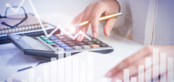 Visão aproximada da mão de uma pessoa usando uma camisa social branca, segurando uma caneta dourada enquanto opera uma calculadora financeira preta sobre uma mesa branca de trabalho. Elementos digitais gráficos e translúcidos estão sobrepostos à cena: uma linha de tendência em ziguezague branca e brilhante aponta para cima, simulando crescimento econômico, acompanhada por um gráfico de barras verticais semitransparente na base da imagem.