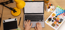 Vista aérea direta (flat lay) de uma mesa de trabalho de madeira organizada. No centro, um notebook está aberto exibindo na tela um wireframe ou esboço digital de design de produto, com mãos digitando no teclado. À esquerda, há uma luminária articulada amarela acesa e uma mesa digitalizadora preta com caneta. À direita, encontram-se um porta-lápis hexagonal branco, um catálogo com estampas geométricas coloridas e uma paleta de cores em leque.