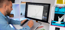 Um designer gráfico de barba curta e camisa jeans trabalha em seu estúdio de criação. Com a mão direita, ele usa uma caneta digital sobre uma mesa digitalizadora cinza para modelar um objeto tridimensional em um monitor grande. A tela exibe um software profissional de modelagem com a malha digital de uma mão humana articulada. À direita, uma impressora 3D de mesa está ligada, fabricando fisicamente a mesma peça plástica da mão modelada na tela. Amostras de cores (paletas) estão organizadas sobre a bancada.