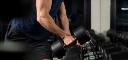 Fotografia em close-up focada no braço musculoso de um homem pegando um halter preto em uma academia. O homem usa uma camiseta regata azul escura e bermuda preta. Ele está segurando o halter pelo centro metálico, levantando-o de um suporte. Abaixo e à direita, há uma longa fileira de halteres idênticos alinhados. O ambiente tem iluminação focada, criando sombras marcadas e um tom geral escuro.
