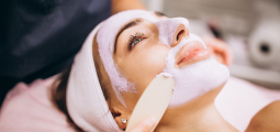 Close-up do rosto de uma mulher deitada de costas com uma faixa branca na cabeça, recebendo um procedimento estético. Uma máscara de argila facial de cor lilás claro está sendo aplicada em suas bochechas e queixo com o auxílio de uma espátula de madeira manejada por um profissional. A cliente está com os olhos abertos e um semblante calmo e relaxado.
