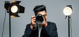 Um jovem fotógrafo de cabelo estilizado e blazer escuro está centralizado contra um fundo cinza de estúdio. Ele segura uma câmera fotográfica profissional DSLR preta na altura do rosto, apontando a grande lente diretamente para a frente, como se estivesse tirando uma foto do observador. Ele está posicionado entre dois refletores de iluminação profissional acesos em tripés pretos, um de cada lado.