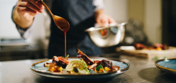 Close-up focado na finalização de um prato gourmet em uma cozinha profissional. Um chef vestindo avental escuro segura uma colher dourada com a mão direita, despejando delicadamente um fio de molho encorpado e avermelhado sobre pedaços de carne grelhada, dispostos artisticamente com purê amarelo e uma folha de repolho tostada em um prato de cerâmica azul. Com a mão esquerda desfocada, ele segura uma tigela de inox.