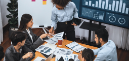 Visão em ângulo superior de uma reunião corporativa ao redor de uma mesa de madeira clara. Uma mulher negra vestindo camisa social azul-clara está em pé na cabeceira, apontando para relatórios com gráficos impressos sobre a mesa. Quatro colegas em trajes de negócios estão sentados ao redor escutando e debatendo. Ao fundo, um painel digital grande exibe gráficos de barras e indicadores comerciais de um dashboard financeiro, ao lado de um quadro branco com notas adesivas coloridas.