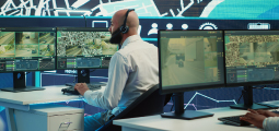 Um operador de segurança de perfil, careca e vestindo camisa social branca com um headset preto na cabeça, trabalha em uma moderna central de monitoramento. Ele está sentado em frente a quatro monitores que exibem transmissões ao vivo de câmeras de segurança (CFTV) com imagens de ruas, rodovias e mapas de satélite. No canto inferior direito, a mão de outro colega digita em um teclado. Ao fundo, há um painel luminoso azul gigante repleto de ícones de tecnologia e gráficos de segurança.