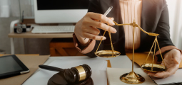 Uma profissional usando paletó preto e blusa plissada em tom pêssego trabalha concentrada em uma mesa de escritório jurídica. Com a mão direita, ela segura uma caneta e ajusta uma tradicional balança da justiça metálica dourada disposta à sua frente. Sobre a mesa, há papéis, um tablet e um martelo de juiz (malhete) de madeira escura com detalhes em latão em primeiro plano. O fundo exibe uma tela de computador e uma câmera fotográfica fora de foco.