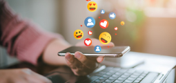 Visão aproximada das mãos de uma pessoa que segura um smartphone na horizontal logo acima de um notebook aberto. Da tela do celular emergem, de forma tridimensional e flutuante, diversos ícones coloridos característicos de redes sociais: corações vermelhos de curtidas, balões azuis com silhuetas de usuários, círculos azuis com o símbolo de positivo ("joinha") e emojis amarelos sorridentes, sendo um deles com corações nos olhos, simbolizando engajamento digital.