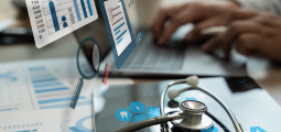 Uma representação conceitual de administração em saúde. Em primeiro plano, um estetoscópio médico preto repousa sobre a tela de um tablet que exibe ícones de dente, DNA e coração. Elementos gráficos digitais translúcidos, como gráficos de barras azuis, relatórios com vistos e uma lupa flutuam sobre a mesa. Ao fundo, as mãos desfocadas de uma pessoa digitam em um notebook.