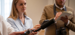 Visão em plano médio de uma entrevista jornalística ou coletiva de imprensa. Em primeiro plano, a extremidade de um microfone de mão preto com espuma protetora contra vento é estendida para registrar uma fala. Logo atrás, fora de foco, uma repórter loira vestindo camisa azul acompanha as declarações com atenção, enquanto um colega de suéter bege anota as informações em um pequeno caderno e traz uma câmera DSLR profissional pendurada no pescoço.