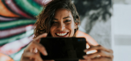 Close-up de uma mulher de cabelos castanhos ondulados segurando um smartphone preto na horizontal bem à frente de seu rosto para gravar um vídeo ou tirar uma foto estilo selfie. Ela exibe um sorriso largo e espontâneo, mostrando os dentes, e usa um pequeno piercing no nariz. O plano de fundo é composto por uma parede decorada com grafites abstratos coloridos em tons de verde, rosa, laranja e preto.