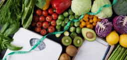 Vista aérea (estilo flat lay) de uma bancada escura repleta de alimentos saudáveis dispostos de forma harmoniosa. Há brócolis, repolho roxo, tomates-cereja, couves-de-bruxelas, pimentões vermelhos e amarelos, abacate, kiwis fatiados, limões e aspargos. Uma fita métrica maleável verde cruza diagonalmente a imagem, passando sobre uma balança mecânica de banheiro branca localizada à esquerda. No canto inferior direito, há uma prancheta de madeira com uma folha de papel em branco e uma caneta azul ao lado.