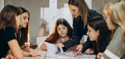 Uma professora de cabelos castanhos lisos e blusa escura está em pé, inclinada com atenção sobre uma mesa de madeira para orientar um grupo de quatro jovens estudantes adolescentes. Ela usa o dedo para apontar e explicar o conteúdo de uma apostila ou livro grande aberto no centro da mesa. As alunas estão sentadas ao redor e olham fixamente para o material com expressões concentradas em uma sala de aula bem iluminada.