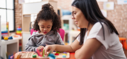 Em uma sala de atendimento infantil com paredes decoradas em tijolos aparentes, uma psicopedagoga de cabelos pretos e óculos está sentada de perfil orientando uma menina negra com cabelos cacheados presos no topo da cabeça. Elas estão juntas em uma pequena mesa vermelha manuseando um tabuleiro pedagógico de madeira que contém blocos e formas geométricas coloridas encachadas. A profissional move delicadamente as mãos para auxiliar a criança na atividade de aprendizado.