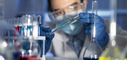 Em um ambiente de laboratório sob iluminação fria e azulada, um cientista focado, usando óculos de proteção transparentes, máscara facial, jaleco branco e luvas de nitrilo azuis, segura um tubo de ensaio de vidro na altura dos olhos. Ele analisa meticulosamente o líquido azul claro contido no tubo. Ao redor e em primeiro plano, suportes de plástico branco seguram diversos outros tubos com substâncias coloridas e balões volumétricos de vidro desfocados.