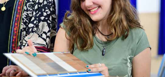 Jovem estudante sorridente, de camiseta verde e cabelo loiro comprido, segura e examina uma paleta de amostras de revestimentos e materiais de acabamento — incluindo tons de madeira e azul — durante uma aula prática. Ao fundo desfocado, outra pessoa e um ambiente de sala de aula colorido.
