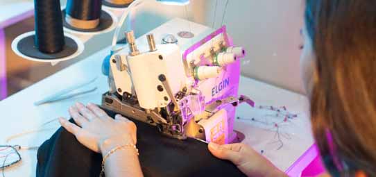 Estudante feminina, de cabelo ruivo solto, opera uma máquina de costura industrial da marca Elgin, costurando tecido preto. A máquina é iluminada por luz roxa/neon, destacando os fios e detalhes do equipamento. Carretéis de linha pretos ao fundo.