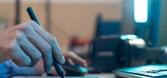 Close em uma mão masculina segurando uma caneta stylus preta sobre uma mesa digitalizadora, com um mouse e teclado ao fundo levemente desfocados. A cena é iluminada por luz azulada de um monitor, sugerindo trabalho em design ou edição gráfica.