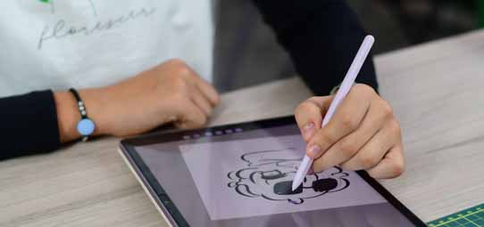 Mão feminina segura uma caneta stylus lilás e desenha diretamente sobre a tela de um tablet, criando uma ilustração digital de personagem em preto e branco com estilo cartoon. Ao fundo, uma régua de corte verde sobre mesa de trabalho.