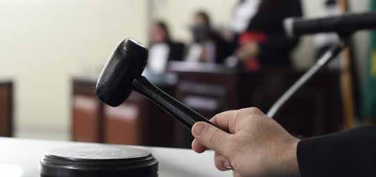 Em primeiro plano nítido, uma mão segura um martelo de juiz (maço) preto sobre sua base circular, em um ambiente de tribunal. Ao fundo desfocado, figuras com roupas formais e togas escuras e vermelhas estão sentadas, compondo o cenário de um júri ou sessão judicial.