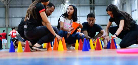 Quatro estudantes de Educação Física agachados em quadra poliesportiva coberta organizam e posicionam cones coloridos — laranja, azul e amarelo — no piso para montar circuito de atividade motora. Uma estudante feminina de camiseta branca estampada segura um cone laranja enquanto os colegas de camisetas pretas trabalham em conjunto. Ao fundo desfocado, outros alunos praticam atividades esportivas na quadra.