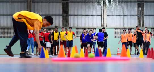 Aula de Educação Física em quadra poliesportiva coberta. Em primeiro plano, um estudante de colete laranja se abaixa para pegar uma bola verde entre cones coloridos (azul, amarelo e vermelho) dispostos no chão. Ao fundo, um grupo numeroso de alunos com coletes coloridos aguarda a atividade com entusiasmo.