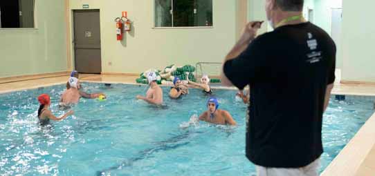 Aula de polo aquático em piscina interna coberta. Um grupo de aproximadamente seis nadadores com toucas coloridas (azul, vermelho e branco) está dentro d'água praticando com uma bola. Um professor de camiseta preta está à beira da piscina, aparentemente orientando a atividade.