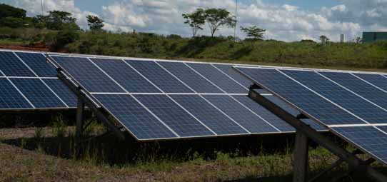 Fileiras de painéis solares fotovoltaicos azuis instalados em estruturas metálicas inclinadas sobre terreno com vegetação rasteira verde. Ao fundo, árvores de médio porte e céu nublado com nuvens. A imagem representa parte de uma usina solar em ambiente rural ou universitário.