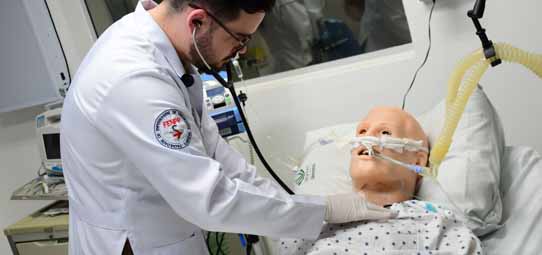 Estudante masculino de Enfermagem, com jaleco branco com brasão de instituição, luvas e estetoscópio, realiza procedimento em manequim de simulação pediátrico intubado deitado em maca hospitalar. O ambiente é um laboratório de simulação clínica equipado com monitores e aparelhos médicos ao fundo.