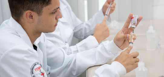 Estudante masculino jovem de Enfermagem, de jaleco branco com brasão institucional vermelho, realiza procedimento de preparo de medicação injetável com concentração. Ele segura um frasco ampola com a mão esquerda enquanto aspira o conteúdo com seringa na mão direita. Ao fundo levemente desfocado, outro estudante realiza o mesmo procedimento. O ambiente claro e organizado remete a laboratório de habilidades clínicas.
