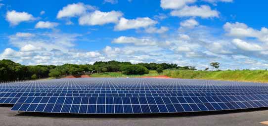 Vista panorâmica de uma grande usina fotovoltaica com centenas de painéis solares azuis instalados em fileiras extensas sobre solo avermelhado. Ao fundo, vegetação verde densa, construções ao longe e céu azul vibrante com nuvens brancas volumosas. A imagem destaca a escala da energia solar renovável.