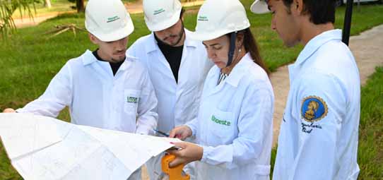 Quatro estudantes de Engenharia Civil usando capacetes brancos e jalecos brancos com logotipo "Unoeste" analisam uma planta ou plano de obras grande estendido. Uma estudante feminina aponta para o documento com uma caneta enquanto os colegas observam. O cenário é externo, em área verde do campus.
