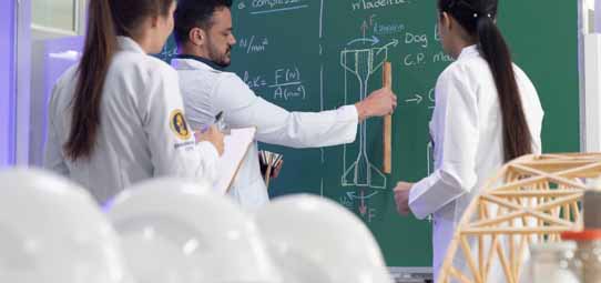 Estudante masculino de jaleco branco aponta para fórmulas e diagramas desenhados em quadro verde — incluindo esboços de estruturas, equações com força e área — enquanto duas estudantes femininas de jaleco, uma com prancheta, observam. Em primeiro plano desfocado, objetos de laboratório e uma estrutura de madeira triangular.