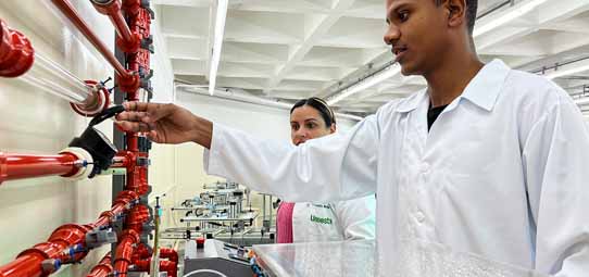 Dois estudantes de jaleco branco — um homem jovem negro e uma mulher — examinam um sistema de tubulações industriais vermelhas montado em bancada de laboratório. O estudante masculino manipula uma válvula enquanto a colega observa. O ambiente é um laboratório de hidráulica ou mecânica de fluidos.