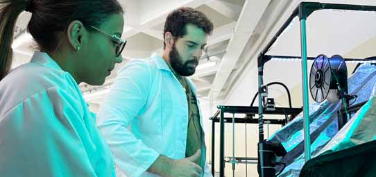 Dois estudantes de jaleco branco — uma mulher de óculos e um homem com barba — observam e operam uma impressora 3D de grande estrutura metálica escura, com carretel de filamento visível na parte superior. A iluminação esverdeada do laboratório cria atmosfera tecnológica.