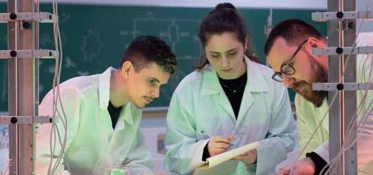 Três estudantes de jaleco branco — dois homens e uma mulher ao centro segurando prancheta — analisam juntos dados ou procedimentos em laboratório iluminado com luz verde. Estruturas de suporte metálicas ao fundo e quadro negro com inscrições compõem o cenário de laboratório de Engenharia Química.