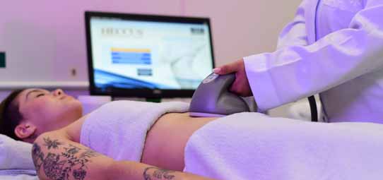 Procedimento estético corporal sendo realizado em cliente feminina deitada em maca, coberta com toalha brancas, com tatuagem floral visível no braço. Mãos de profissional de jaleco branco aplicam aparelho de ultrassom ou radiofrequência sobre o abdômen da paciente. Ao fundo, monitor de computador exibe interface de equipamento estético com parâmetros de tratamento. Iluminação roxa suave cria atmosfera de clínica de estética.