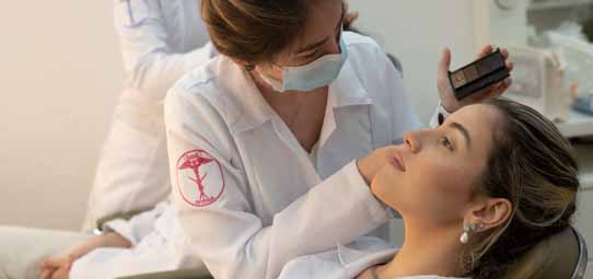 Estudante de Estética e Cosmética, com jaleco branco com logotipo rosa de figura feminina e máscara facial, realiza procedimento estético facial em paciente reclinada em maca de atendimento. A profissional segura um equipamento de aplicação — aparentemente caneta de microagulhamento ou radiofrequência — e trabalha na região da testa da cliente. A expressão da paciente é tranquila e relaxada. Ambiente clínico claro ao fundo.